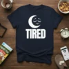 Navy t-shirt with white text "TIRED" and a sad, sleepy moon graphic above it