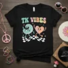 Black t-shirt with "TK VIBES" text and retro cartoon characters, set against a checkered pattern