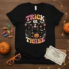 Trick Or THREE Halloween t-shirt with a cute black cat holding a pumpkin, surrounded by flowers and bats