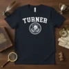 Navy t-shirt with white "TURNER" text in varsity font above a shield with a lion and laurel wreath