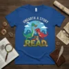 Blue t-shirt with cartoon dinosaurs reading books, text says "Unearth a Story Read"