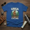 Ursa Cycling t-shirt design featuring a cartoon bear wearing sunglasses and a helmet, riding a bicycle through a forest. Text