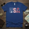 Blue t-shirt with bold "USA" text; the "U" features the Statue of Liberty, the "S" is a US flag pattern
