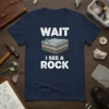 Navy blue t-shirt with white text "WAIT I SEE A ROCK" and a graphic of rock layers with a pickaxe