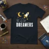 We are all DREAMERS t-shirt with a rocket, moon, and stars graphic in yellow and white on a navy blue