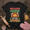 Black t-shirt with "WE HAVE COOKIE IN THE OVEN" text and gingerbread men in an oven graphic, festive holiday theme