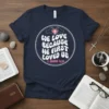Christian t-shirt with white text "We Love Because He First Loved Us" and "1 John 4:19" below, featuring hands forming