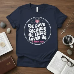 Christian t-shirt with white text "We Love Because He First Loved Us" and "1 John 4:19" below, featuring hands forming