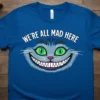 Blue t-shirt with the text "We're All Mad Here" above a grinning Cheshire Cat face, featuring bright green eyes and