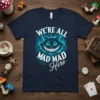 T-shirt with "WE'RE ALL MAD MAD Here" text and a smiling Cheshire Cat graphic in teal and white