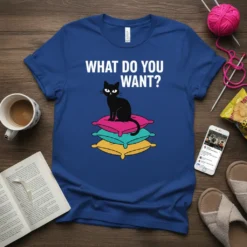 Black cat sitting on stacked colorful pillows with "WHAT DO YOU WANT?" text above, on a royal blue t-shirt