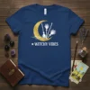 Navy blue t-shirt with white text "Witchy Vibes" and a golden crescent moon graphic with white broomsticks