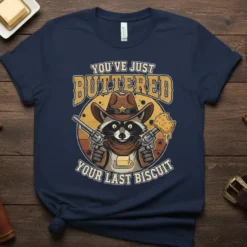 Funny cowboy raccoon holding guns and buttered biscuit, text "You've Just Buttered Your Last Biscuit" on navy t-shirt