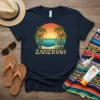Zanzibar text with a tropical beach sunset design featuring palm trees, ocean, and surfboard graphic on a navy t-shirt