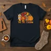 Graphic t-shirt design featuring the text 'Tis the season with autumn leaves, a football, and a pumpkin